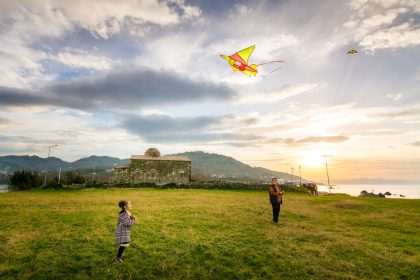 Uma criança e um adulto empinam pipas em uma paisagem rural ao pôr do sol.