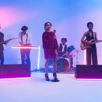 Banda se apresenta em um palco iluminado por luzes coloridas, com instrumentos musicais ao fundo.