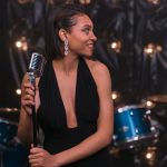 Cantora se apresenta ao microfone em um palco, com uma bateria ao fundo.