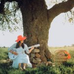Mulher usando chapéu vermelho toca violão acústico sentada sob uma árvore.