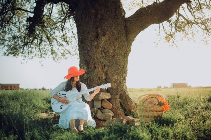 Mulher usando chapéu vermelho toca violão acústico sentada sob uma árvore.
