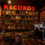 Interior de uma loja de discos com prateleiras de vinis, decoração retrô e letreiro luminoso escrito “Records”.