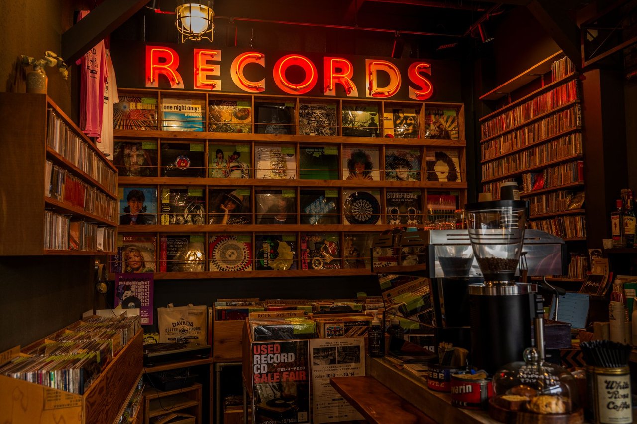 Interior de uma loja de discos com prateleiras de vinis, decoração retrô e letreiro luminoso escrito “Records”.