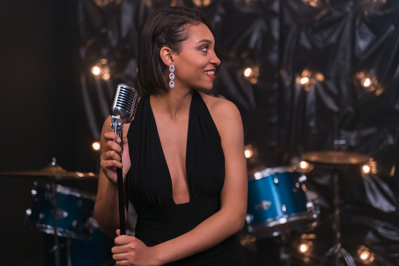 Cantora se apresenta ao microfone em um palco, com uma bateria ao fundo.