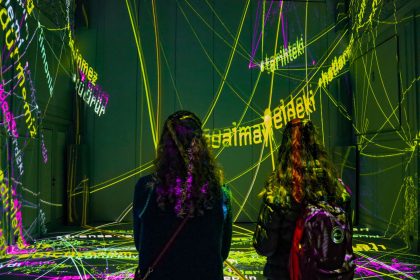 Duas mulheres observam uma instalação artística com projeção de textos em uma sala escura.
