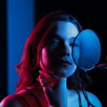 Cantora grava vocais diante de um microfone em um estúdio iluminado com luzes coloridas.